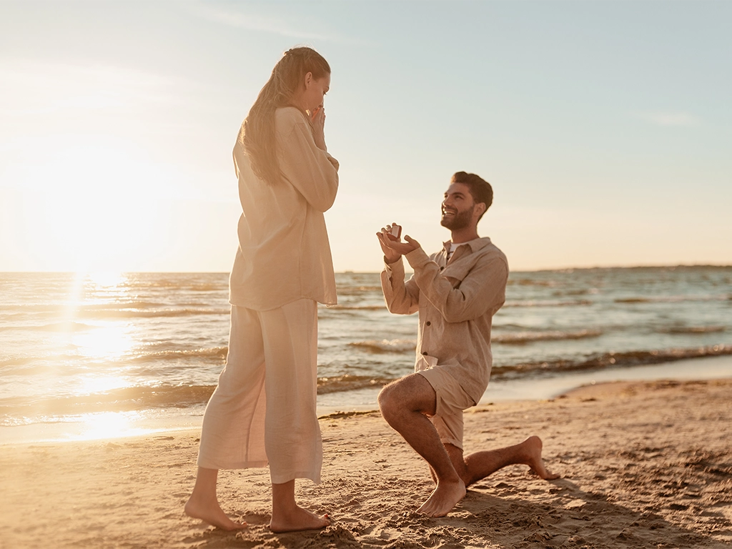 smiling young man with engagement ring making proposal to happy woman on beach